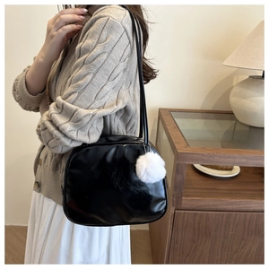 Detail of a black glossy shoulder bag featuring a white fluffy pompom charm and zipper