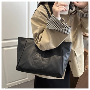 Close-up of black lychee-textured tote bag with circular embossed logo on khaki coat