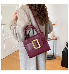 Model wearing a wine red crossbody bag with a large gold buckle and top handle over a coat