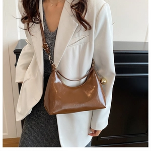 Detail of a brown patent leather crescent bag featuring a gold ball charm and wide strap