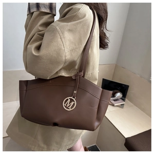 Detail of dark brown tote bag with metal M charm on a model in a beige trench coat