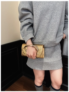 Detail of a gold textured clutch bag featuring a large oval metal handle on a grey outfit