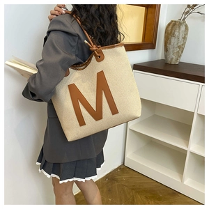 Detail of a large beige woven tote bag with a brown leather M patch worn by a model