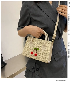 Detail of woven straw handbag featuring 3D red cherry accents and a neutral shoulder strap