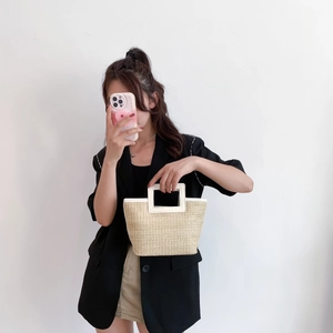 Detail of woven straw tote bag with white leather handle held by model in black blazer