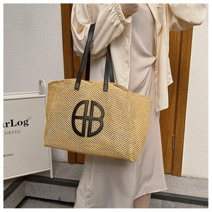 Close-up of woven straw tote bag with black HB logo and leather shoulder strap on a model