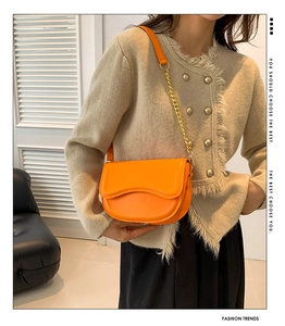 Detail of orange saddle bag featuring wave-shaped flap and gold metal chain strap