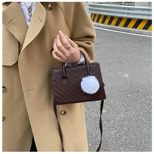 Detail view of dark brown quilted small square bag with white fur ball charm and shoulder strap