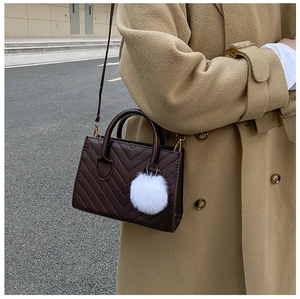 Detail of dark brown V-quilted handbag with a white fur pompom on a camel wool coat background