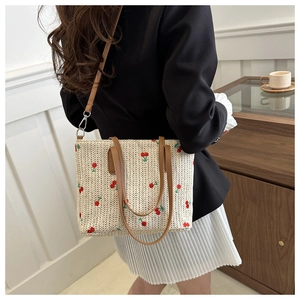 Model wearing a woven straw tote bag with red cherry embroidery and brown leather handles