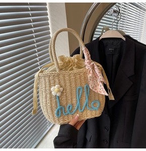Detail of hand-woven straw tote bag with blue hello text, daisy charm and floral scarf
