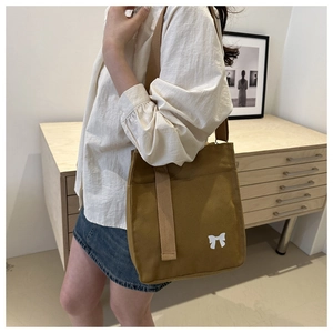 Detail of a brown canvas shoulder bag with white bow logo worn by a model in a cozy room
