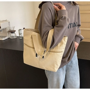 Model wearing a large beige canvas tote bag with front zip pockets and woven straps