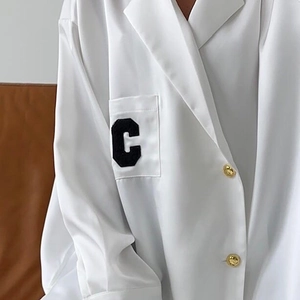Detail der weißen Satinbluse mit geprägten Goldknöpfen und schwarzem Buchstaben-C-Patch