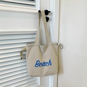 Minimalistische beige Tragetasche mit blauem Beach-Schriftzug an weißer Wand