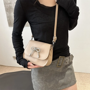 Elegant beige saddle bag with rhinestone bow detail shown on model in minimalist setting