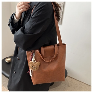 Brown faux leather tote bag with plush bear charm worn by a model in a minimal room