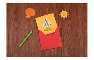 Close-up of a yellow 3D Christmas card with a green tree and red envelope on a wooden desk