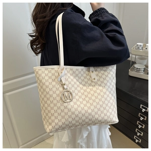 Detail of a large beige tote bag with M-pattern and gold pendant on a woman's shoulder