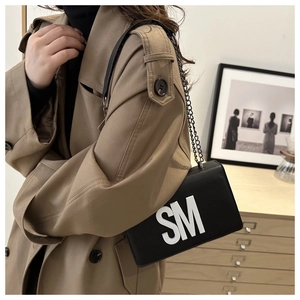 Detail of a minimalist black leather chain bag with 3D white SM letters on a trench coat