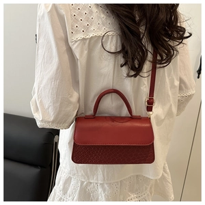 Close-up of red crossbody bag with woven texture and top handle on a white lace shirt