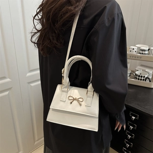 White faux leather handbag with a gold bow buckle worn by a model in a modern interior