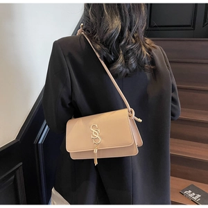 Beige crossbody bag with gold S-logo and fringe tassel shown on model in minimalist setting