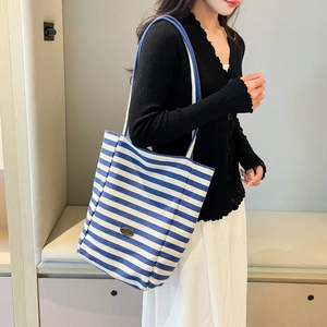 Blue and white striped canvas tote bag worn by a model in a minimalist interior setting
