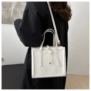 Vertical square white crossbody bag shown on shoulder, featuring dual handles and metal tassel