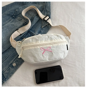 Flat lay of a white waist bag featuring pink bow embroidery with a denim jacket and phone