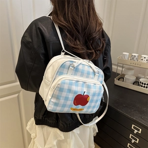 Blue and white plaid backpack with red apple print worn by a model in a cozy room