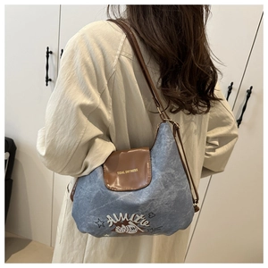 Detail of denim shoulder bag featuring cartoon embroidery and brown leather flap with gold text