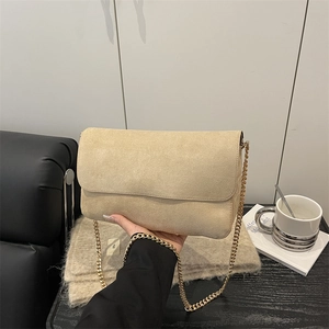 Minimalist beige suede shoulder bag with gold chain strap shown in a cozy cafe setting