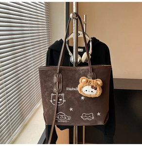 Detail van een grote bruine tote bag met wit kattenborduurwerk en een pluchen beerhanger