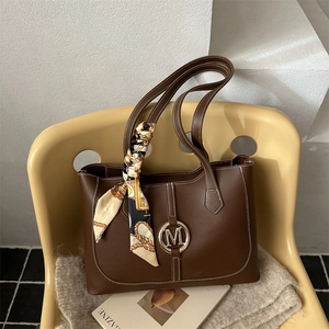Bruine lederen shopper met gouden M-logo en decoratieve zijden sjaal op hippe stoel