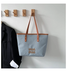 Detail of light blue woven tote bag with brown leather handles and brand logo on white wall