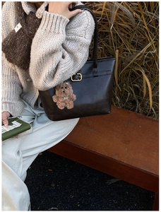 Detail of dark brown leather tote bag with gold buckle and cute bear charm on a park bench