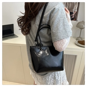 Detail of a black glossy leather tote bag with slim straps worn by a model in a modern room