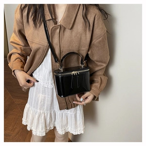 Modern black patent leather box handbag with gold hardware shown on person with brown coat