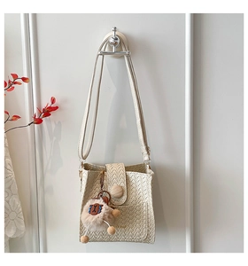 Detail of cream woven crossbody bag with gold logo and plush charm on white wall