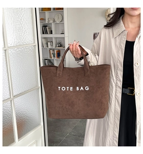 Detail of a brown suede tote bag with white text logo held by a person in a minimalist room