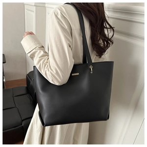 Black large tote bag with gold metal plaque worn by a model in a minimalist room