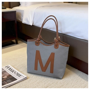 Detail of grey woven tote bag featuring large brown M patch and leather piping accents