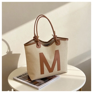 Detail of beige straw tote bag with brown leather handles and a large letter M patch