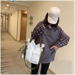 White canvas tote bag with green straps and dinosaur charm shown on a model in a hallway