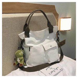 White canvas tote bag featuring double front pockets and a green dinosaur plush charm