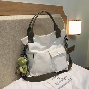 Cream canvas tote bag with green straps and cute dinosaur plush charm, shown on a bed