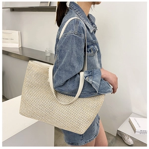 Large woven straw tote bag with white leather straps worn by a model in casual style