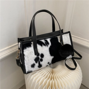 Faux fur cow print handbag with black leather straps and plush ball charm on white stand