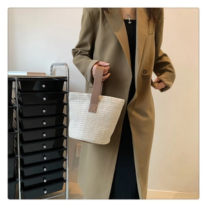 Detail of straw woven bucket bag with brown wide handles held by a model in a trench coat